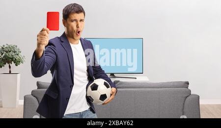 Jeune homme décontracté tenant un ballon de football et montrant un carton rouge devant la télévision isolé sur fond blanc Banque D'Images