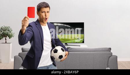 Jeune homme tenant un ballon de football et montrant un carton rouge devant la télévision isolé sur fond blanc Banque D'Images