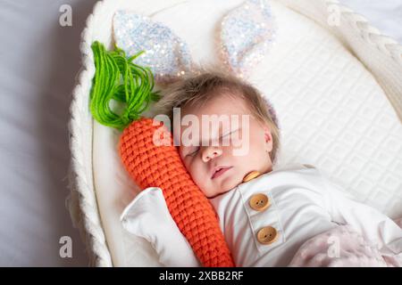 Petit bébé nouveau-né dormant dans un berceau. Bébé dormant dans un panier Moïse, portant des oreilles de lapin scintillantes et tenant une carotte. Banque D'Images