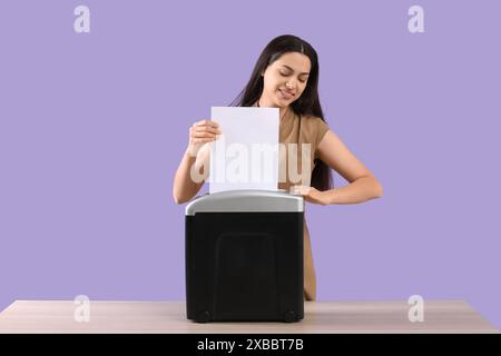 Jeune femme détruisant la feuille de papier à l'aide d'un déchiqueteur sur la table sur fond lilas Banque D'Images