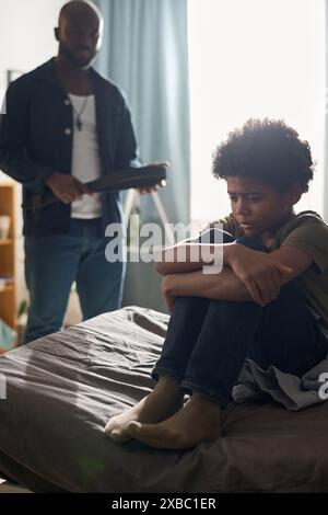 Portrait vertical pleine longueur du jeune garçon noir pleurant assis sur le lit avec parent en colère tenant la ceinture en arrière-plan Banque D'Images