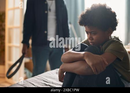 Vue de côté portrait de petit garçon noir pleurant assis sur le lit dans l'obscurité avec père en colère tenant la ceinture pour l'espace de copie de punition Banque D'Images