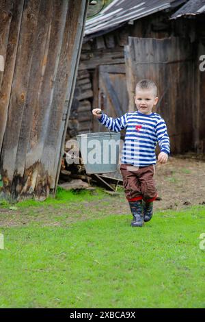 Un garçon portant un seau en métal, portant une chemise rayée, un pantalon brun et des bottes en caoutchouc, marchant sur l'herbe dans un cadre rustique et rural.Belarus.Minsk Banque D'Images