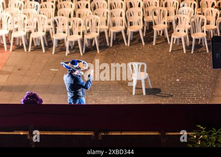 Un chanteur dans un sombrero se produisant sur une scène extérieure vide avec des chaises blanches en arrière-plan. Capturer l'essence d'une performance solitaire. Banque D'Images