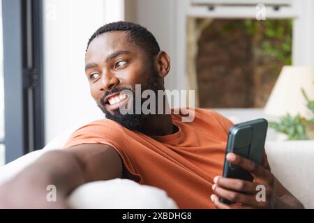 Jeune homme afro-américain tenant un smartphone, souriant tout en se relaxant sur le canapé, à la maison. Salon lumineux avec grande fenêtre montrant la verdure à l'extérieur Banque D'Images
