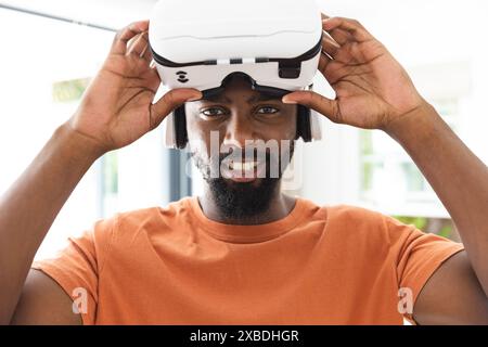 Jeune homme afro-américain portant un casque de réalité virtuelle, souriant à la caméra. Cadre intérieur lumineux avec grandes fenêtres et lumière naturelle Banque D'Images