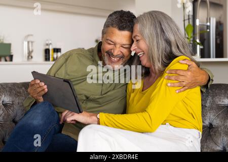 Un couple biracial mature est assis sur un canapé, souriant et regardant une tablette, à la maison Banque D'Images