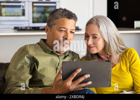 Un couple biracial mature regarde une tablette ensemble, à la maison. Ils sont assis sur un canapé dans une cuisine moderne, profitant d'un moment calme à la maison, una Banque D'Images
