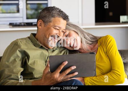Un couple biracial mature sourit en regardant une tablette ensemble, à la maison. Ils sont assis sur un canapé confortable dans un salon moderne avec un Banque D'Images