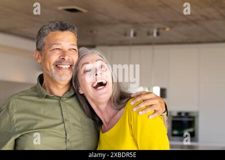 Un couple biracial mature sourit chaleureusement à la maison, à la maison. Ils sont debout dans une cuisine moderne avec des plafonds en bois et des armoires blanches, inchangées Banque D'Images