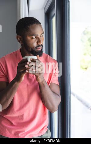 Jeune homme afro-américain tenant une tasse de café, regardant soigneusement par une fenêtre, à la maison. Une lumière naturelle éclatante illumine l'intérieur d'une maison moderne avec un Banque D'Images
