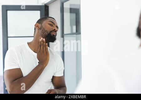 Jeune homme afro-américain toilettant la barbe devant le miroir, à la maison. Salle de bains moderne avec murs de fond blanc et porte vitrée, créant élégant et Banque D'Images