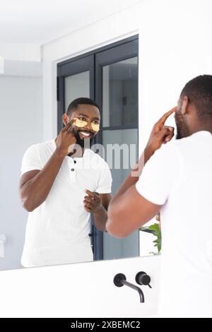 Jeune homme afro-américain appliquant des soins de la peau devant le miroir de la salle de bain, à la maison. Salle de bains lumineuse et moderne avec grand miroir et fenêtre en arrière-plan, Banque D'Images