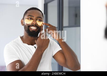 Jeune homme afro-américain appliquant des patchs sous les yeux, souriant à son reflet, à la maison. Salle de bains moderne avec murs blancs et grandes fenêtres, naturel Banque D'Images