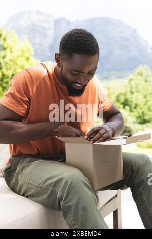 Jeune homme afro-américain ouvrant le paquet tout en étant assis à l'extérieur, à la maison. Verdure luxuriante et montagnes créant un arrière-plan serein, ajoutant à la paix Banque D'Images