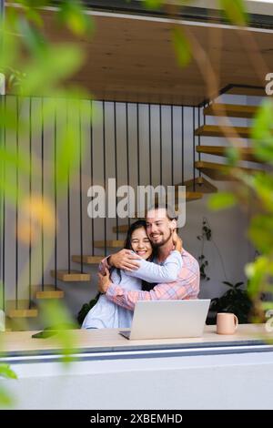 Jeune couple diversifié embrasse tout en étant assis à une table avec un ordinateur portable Banque D'Images