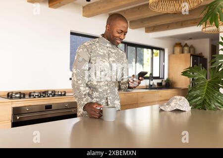 Jeune soldat afro-américain en uniforme militaire tient une tasse et regarde son smartphone. Cuisine moderne avec des accents en bois et de grandes fenêtres à l'arrière Banque D'Images