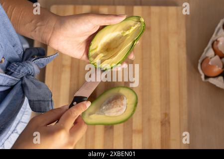 Jeune femme biraciale coupant l'avocat dans une cuisine confortable. Planche à découper en bois et coquilles d'œufs sur le comptoir, avec une atmosphère chaleureuse améliorant la scène, unalt Banque D'Images