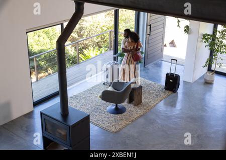 Jeune couple diversifié se tient avec des bagages dans une maison de vacances moderne. De grandes fenêtres révèlent une végétation luxuriante à l'extérieur, créant une atmosphère sereine et accueillante Banque D'Images