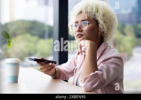 Jeune femme biraciale tenant smartphone, regardant réfléchi dans le bureau d'affaires. fenêtre montrant le paysage urbain et la verdure à l'extérieur, mélangeant urbain et natu Banque D'Images