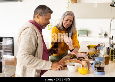 Un couple biracial mature prépare le repas ensemble dans une cuisine lumineuse, à la maison. Le comptoir rempli d'ingrédients frais et d'articles de petit déjeuner crée un Banque D'Images