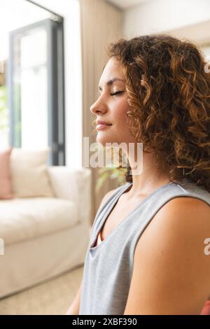 Jeune femme caucasienne se relaxant les yeux fermés, méditant. Espace de vie intérieur avec mobilier de couleur claire et lumière naturelle, inaltérable Banque D'Images