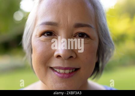 Femme senior asiatique souriant chaleureusement avec les cheveux gris courts, à l'extérieur, à la maison. À l'extérieur dans un jardin luxuriant avec la lumière douce du soleil filtrant à travers les arbres, créatine Banque D'Images