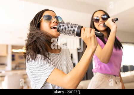 Deux jeunes sœurs biraciales chantent dans des brosses à cheveux tout en portant des lunettes de soleil, à la maison Banque D'Images