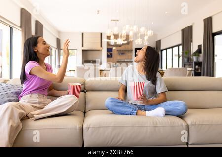 Deux jeunes sœurs biraciales sont assises sur un canapé, dégustant du pop-corn et riant ensemble, à la maison. Ils sont dans un salon moderne avec de grandes fenêtres et St Banque D'Images