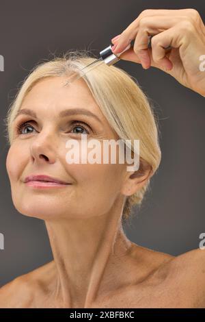 Femme Jolly mature appliquant le sérum sur une toile de fond grise. Banque D'Images