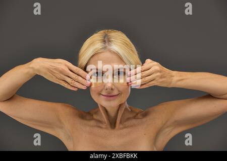 Charmante femme mature avec des cheveux blonds avec des patchs pour les yeux sur un fond gris. Banque D'Images