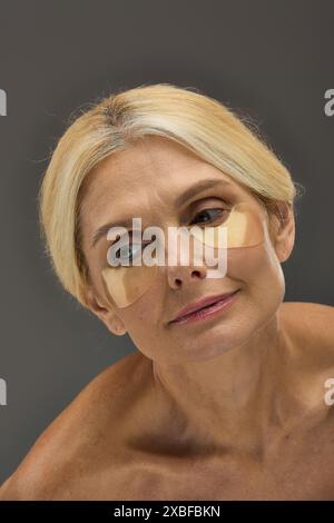 Femme mature attrayante avec des cheveux blonds avec des patchs oculaires sur un fond gris. Banque D'Images