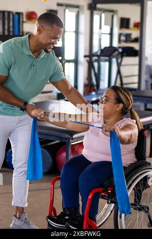 Femme biraciale quadriplégique en fauteuil roulant utilisant une bande de résistance, assistée par un homme. Ils sont dans le centre de réadaptation avec équipement d'exercice et therap Banque D'Images
