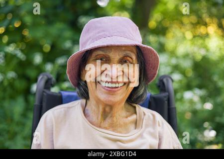 Vieille femme indienne (sud-asiatique) dans ses années 90 souriante regardant la caméra, portant des vêtements décontractés et un chapeau assis dans un fauteuil roulant à l'extérieur en été, U. Banque D'Images