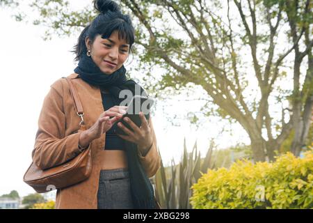 Une femme latine heureuse des notifications sur son téléphone portable. Elle porte une veste marron et une écharpe noire, défilant et tapant sur l'écran pendant Banque D'Images