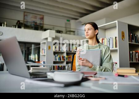 Une adolescente profondément dans la pensée alors qu'elle travaille sur ses devoirs à l'aide d'un ordinateur portable dans un cadre de bibliothèque calme. Banque D'Images