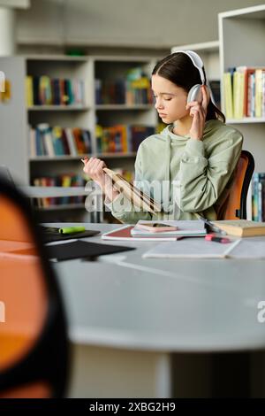 Une adolescente, portant des écouteurs, se concentre sur son ordinateur portable alors qu'elle est assise à un bureau dans une bibliothèque. Banque D'Images