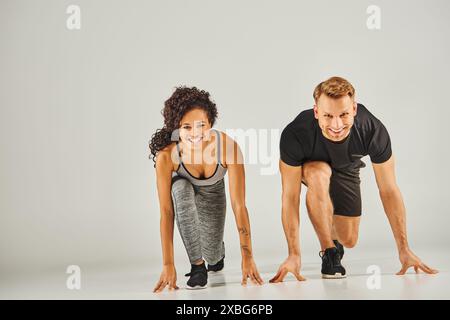 Jeune couple de sport interracial en vêtements actifs poussant ensemble sur fond gris, mettant en valeur la force et le travail d'équipe. Banque D'Images