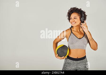 Femme afro-américaine aux cheveux bouclés en vêtements de sport, portant des écouteurs, tenant une balle, Banque D'Images
