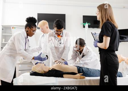 Quatre étudiants en médecine en manteau blanc pratiquent la RCR sur un mannequin dans une salle de classe. Une femme vêtue de noir observe la séance d'entraînement Banque D'Images