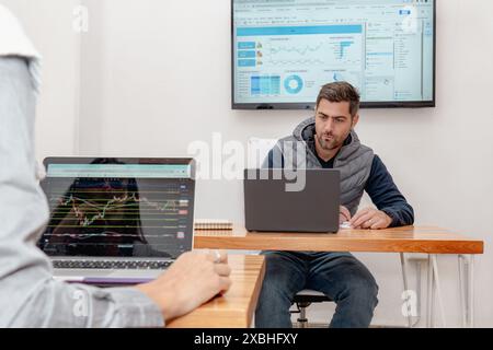 Un homme d'âge moyen est profondément concentré sur son ordinateur portable dans un espace de coworking moderne, avec un écran affichant des graphiques derrière lui. Une autre personne travaille à proximité Banque D'Images