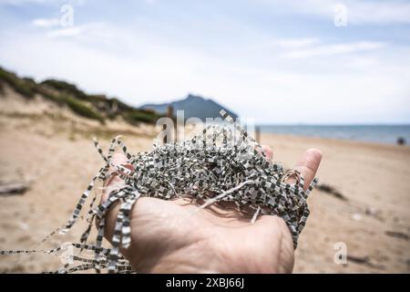 Main tenant des débris en plastique sur une plage de sable avec la mer en arrière-plan. Les rubans proviennent de la désintégration de sacs de raphia synthétiques. Un concp Banque D'Images