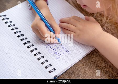 Écrire avant l'école, la fille d'âge préscolaire dessine des éléments avec un stylo dans un cahier à carreaux, développement physique de la main, premiers pas dans l'alphabétisation Banque D'Images