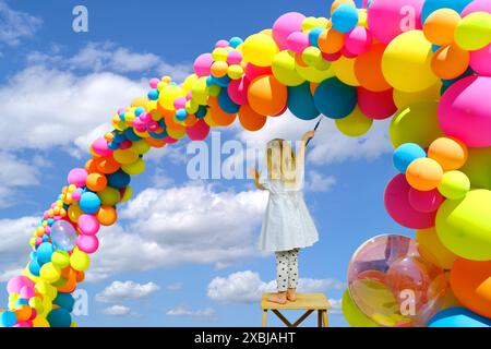 petit enfant, fille blonde 2 ans en robe blanche vieux se tient sur chaise haute et dessine dans l'arc-en-ciel coloré du ciel, ballons de vacances couleurs vives, concept de Banque D'Images