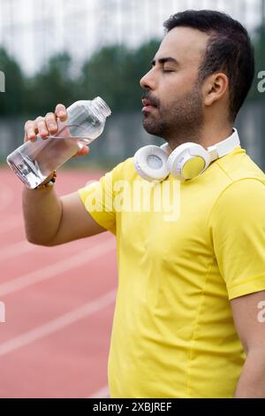 Le sportif hydratant dans les écouteurs sans fil prend une gorgée rafraîchissante de la bouteille d'eau sur la piste de course Banque D'Images