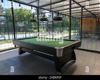 Une table verte de billard se trouve dans une aire de loisirs extérieure couverte, avec un terrain de basket-ball et des tables de tennis visibles autour. Banque D'Images