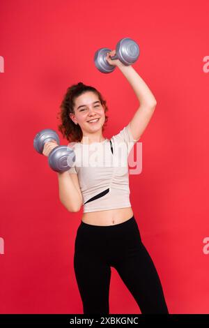 Adolescente sportive exerce avec des haltères pour développer des muscles isolés sur fond rouge. Banque D'Images