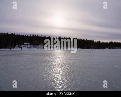 Vue de paysage de lac gelé avec des arbres à l'arrière et réflexion de lumière sous le ciel d'humeur changeante Banque D'Images