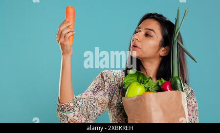 Femme avec un sac en papier dans les mains rempli de légumes vérifiant la carotte pour les impuretés. Client vivant conscient avec des produits d'épicerie achetés à utiliser comme ingrédients de cuisine, fond de studio, caméra B. Banque D'Images