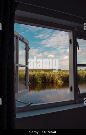 Une fenêtre ouverte donnant sur un paysage tranquille avec un canal, des prairies et un ciel nuageux, Ilpendam, pays-Bas Banque D'Images